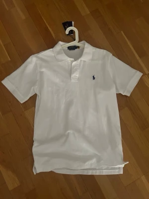    Polo Ralph Lauren pikétröja - Klassisk vit pikétröja från Polo Ralph Lauren med blå broderad logga på bröstet. Tröjan har korta ärmar, krage och knäppning framtill. Perfekt basic till garderoben och enkel att matcha med allt. Helt ny, inga skador. Aldrig använd, säljer för att den är för stor för mig. 