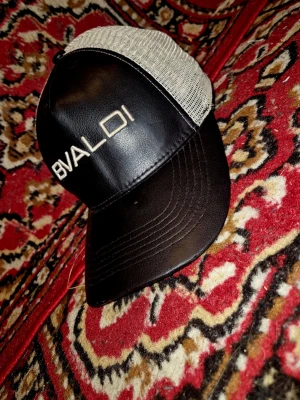 Svart och beige keps från BVALDI - Snygg keps från BVALDI med svart framsida i läderlook och vit mesh baktill. Broderad logga framtill i vitt. Perfekt för en cool och avslappnad stil.