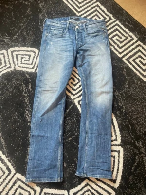 Blå raka jeans med fem fickor - Säljer ett par klassiska blå jeans med raka ben och fem fickor. Jeansen har en ljus tvätt och subtila slitningar för en avslappnad look. Passar perfekt till en casual stil och har knapp- och dragkedjestängning.