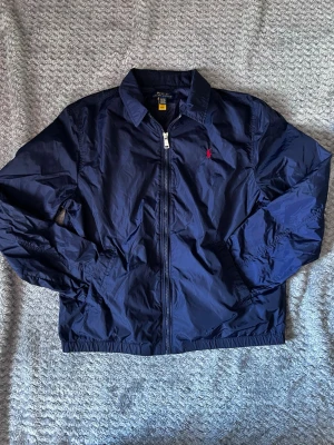 Marinblå vindjacka från Polo Ralph Lauren - Snygg marinblå vindjacka från Polo Ralph Lauren med dragkedja framtill och klassisk krage. Jackan har röd broderad logga på bröstet och är tillverkad i lätt, glansigt material. Perfekt för blåsiga dagar och enkel att matcha med olika outfits.