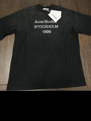 Svart Acne Studios t-shirt med tryck - Svart t-shirt från Acne Studios med vit text 'Acne Studios STOCKHOLM 1996' på bröstet. Klassisk passform med rund hals och korta ärmar. Tillverkad i mjuk bomull, perfekt för en stilren och avslappnad look.