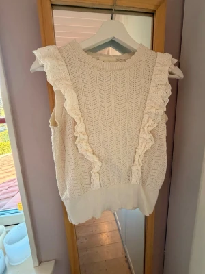 Vit ärmlös stickad topp med volang - Supersöt vit ärmlös stickad topp med volangdetaljer framtill och runt axlarna. Toppen har ett fint hålmönster och bred ribbad kant nedtill. Perfekt att styla med jeans eller kjol för en trendig look.