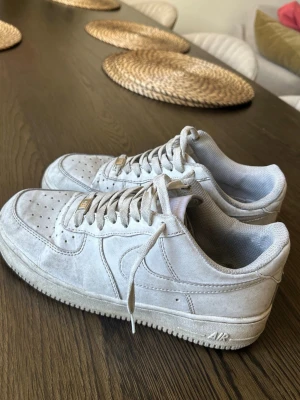 Nike Air Force 1 vita mockasneakers - Snygga Nike Air Force 1 sneakers i vitt mockaläder med klassisk siluett och perforerad tå. Skorna har låga skaft, vita skosnören och diskret Nike-logga på sidan. Perfekta för en clean och stilren look.