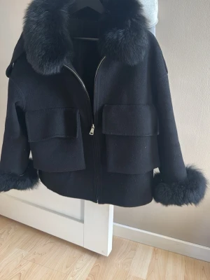 Svart pälsjacka med stora fickor - Snygg svart pälsjacka med fluffig rävpäls vid krage och ärmslut. Jackan har två stora fickor framtill och dragkedja. Ger en lyxig känsla med avtagbar päls till våren. Materialet är tjockt och värmande. Från ssshop nypris: 3899kr