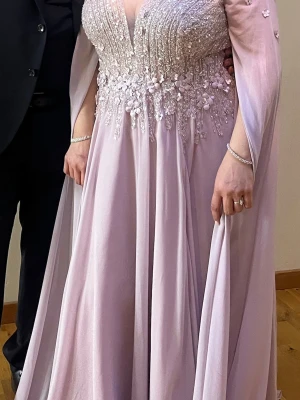 Ljuslila långklänning med pärldetaljer - Elegant ljuslila långklänning med v-ringning och långa, transparenta ärmar. Överdelen är dekorerad med pärlor och blommiga applikationer som ger en lyxig känsla. Kjolen är svepande och ger ett vackert fall. Perfekt för bal eller fest. Storlek L passar LX