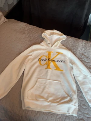 Vit hoodie från Calvin Klein Jeans - Snygg vit hoodie med stor gul CK-logga och svart text på bröstet. Klassisk känguruficka framtill och huva med snörning. Perfekt för en avslappnad och trendig stil.