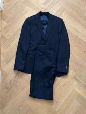 Mörkblå kostym med kavaj och byxor - Snygg mörkblå kostym med klassisk kavaj och matchande byxor. Kavajen har två knappar, fickor och slät finish. Byxorna har pressveck och bakficka med knapp. Perfekt för dig som vill ha en stilren och tidlös look.