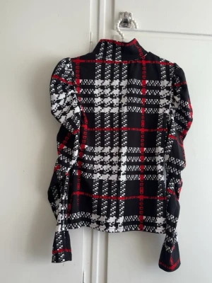 Rutig tröja svart, vitt och rött - Snygg tröja med rutigt mönster i svart, vitt och rött. Skjortan har långa ärmar och en klassisk krage. Materialet känns mjukt och lätt, perfekt för en trendig look.