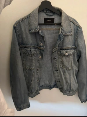 Ljusblå jeansjacka från Bik Bok - Klassisk ljusblå jeansjacka från Bik Bok med två bröstfickor med lock och knappar, samt knappstängning framtill. Jackan har en rak passform och är tillverkad i denim. Perfekt för en avslappnad och trendig look. Saknar en knapp vid fickan vid bröstet 