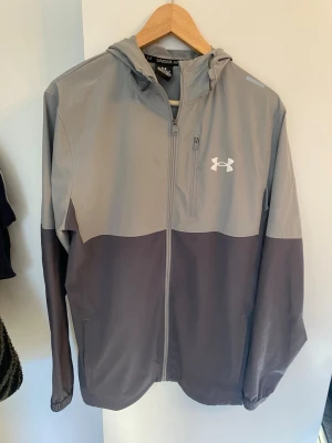 Grå vindjacka från Under Armour - Snygg tvåfärgad vindjacka från Under Armour i ljusgrått och mörkgrått. Jackan har huva, dragkedja framtill, bröstficka med dragkedja och elastiska muddar. Perfekt för blåsiga dagar och sportiga outfits.