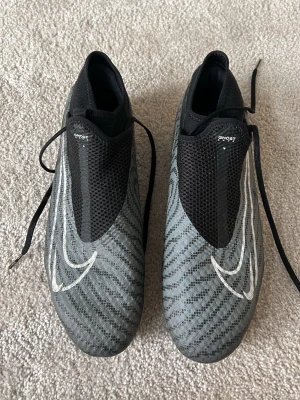 Nike Phantom Vision Ghost fotbollsskor - Snygga gråa Nike Phantom Vision Ghost fotbollsskor med svart mesh och vita detaljer. Skorna har en strömlinjeformad design, snörning på sidan och mönstrad ovandel för bättre bollkontroll. Yttersulan har fasta dobbar för spel på gräs.