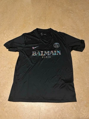 Svart Balmain x PSG t-shirt - Snygg svart t-shirt från Balmain x PSG i samarbete med Nike. T-shirten har korta ärmar, rund hals och är gjord i ett glansigt syntetmaterial. Framtill finns Balmain Paris-tryck och PSG-logga samt Nike swoosh i regnbågseffekt.