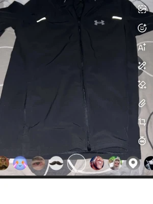 Svart vindjacka från Under Armour - Svart vindjacka från Under Armour med hel dragkedja, huva och reflekterande detaljer på bröstet och ryggen. Jackan har logga på bröstet och stor text på ryggen. Perfekt för löpning eller utomhusaktiviteter.