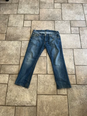 True religion - Säljer ett par klassiska blå jeans med snygga slitningar och tvättade detaljer. Jeansen har fem fickor, raka ben och knappstängning. Perfekt för dig som gillar en avslappnad och tidlös look. Obs litet hål i ena bakfickan
