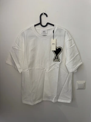 Vit t-shirt från Ami med hjärtlogo - Säljer en vit t-shirt från Ami med en stor svart hjärtformad logo på bröstet. T-shirten har klassisk rund hals och korta ärmar. Materialet är mjuk bomull och passformen är loose. Perfekt för dig som gillar stilrena plagg med coola detaljer.