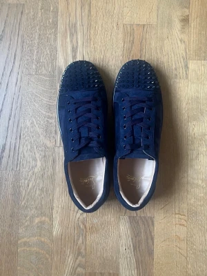 Mörkblå sneakers från Christian Louboutin - Snygga mörkblå sneakers från Christian Louboutin med spetsig tå och coola nitar på tån. Skorna är gjorda i mocka och har klassiska röda sulor. Perfekt för dig som vill sticka ut med ett exklusivt märke och unik design.