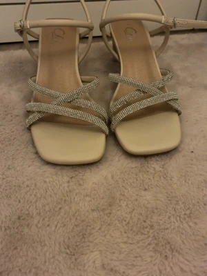 Beige sandaler med strass och klack - Säljer ett par snygga beige sandaler från Call it Spring med fyrkantig tå, medelklack och tunna remmar dekorerade med glittrande strass. Remmarna korsas över foten och har justerbart spänne vid ankeln. Perfekta för att lyfta din outfit. Köpta ifrån skopukten för 449kr men säljer dom för 359kr pris går alltid att diskutera jag har använt dom en enda gång och inte mer dom är i storlek 36.🤗