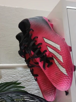 Adidas X rosa och svarta fotbollsskor - Snygga Adidas X fotbollsskor i rosa och svart med vita ränder på sidan. Skorna har snörning, låg profil och greppvänliga dobbar. Ovandelen är tillverkad i ett syntetmaterial som ger bra bollkontroll och lätt känsla på planen.