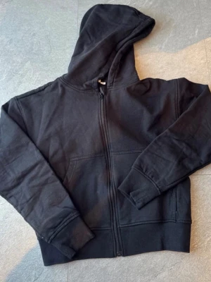 Svart zip-hoodie med huva - En klassisk svart hoodie med dragkedja och huva. Hoodien har ribbade muddar vid ärmslut och nederkant samt en stor magficka framtill. Perfekt för en avslappnad stil. Materialet känns mjukt och bekvämt, troligen bomullsblandning.