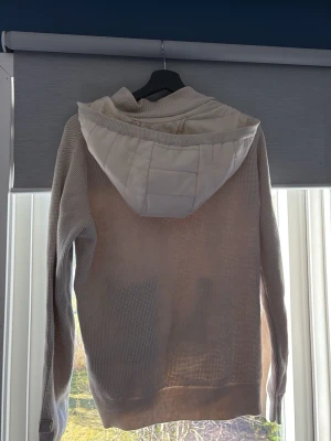 Beige hoodie med dragkedja och huva - Snygg beige hoodie med dragkedja framtill och två fickor med dragkedja. Huvan är vadderad och ger en cool kontrast till resten av tröjan som är ribbstickad. Perfekt för en avslappnad stil och enkel att matcha med jeans eller joggers.