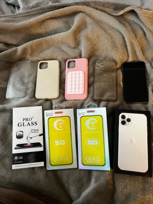 iPhone 11 Pro Silver med tillbehör - Säljer en silverfärgad iPhone 11 Pro med tre kameror. Medföljer flera skal i olika färger och material, inklusive ett rosa pop-it skal, samt tre skärmskydd i glas. Mobilen är väl omhändertagen men har repor på farmskärmen, reporna syns bara i vissa ljus men man blir inte störd av de och en kantstöttning på ena bak kameran. Allt funkar som det ska, mobilen är i fint skick överlag. Skriv gärna vid frågor 😊