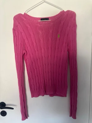 Rosa kabelstickad tröja från Ralph Lauren - Säljer en rosa kabelstickad tröja från Ralph Lauren med den klassiska gröna loggan broderad på bröstet. Tröjan har rund halsringning, långa ärmar och ribbade muddar. Perfekt för dig som gillar färg och stilren design.