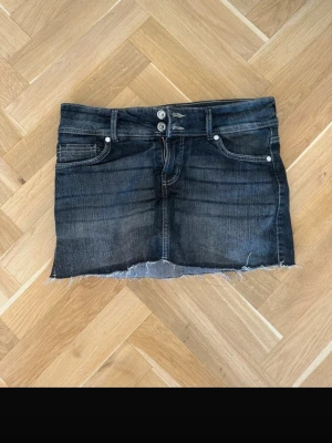 Svart jeanskjol med inbyggda shorts - Cool svart jeanskjol med rå, fransig kant nertill och klassiska fem fickor. Kjolen har dubbla knappar i midjan och dragkedja. Inbyggda shorts