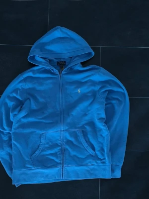 Blå hoodie från Polo Ralph Lauren - VANLIG STORLEK S - BARN STORLEK 170/XL       NYPRIS 2500KR                                                              Snygg blå hoodie från Polo Ralph Lauren med dragkedja och huva. Klassisk gul logga broderad på bröstet och två fickor framtill. Perfekt för chill dagar eller när du vill ha en enkel men stilren look.