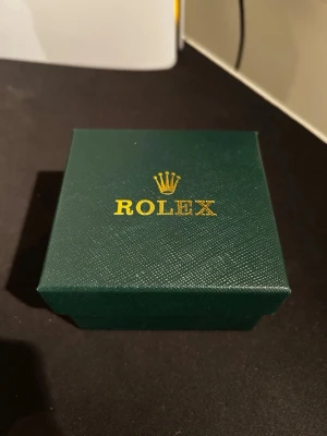 Rolx  - Säljer en Rolx Submariner med grön urtavla och guldfärgade detaljer. Klockan har en klassisk rund form, guldfärgad länk och grönt vridbart bezel. Kommer i original grön Rolxbox. Perfekt för dig som vill ha en ikonisk och lyxig accessoar.