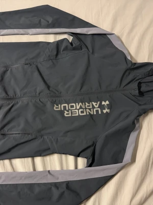 Svart vindjacka från Under Armour - Snygg svart vindjacka från Under Armour med gråa detaljer längs ärmarna och stort logotryck på bröstet. Jackan har dragkedja framtill och huva, perfekt för blåsiga dagar. Materialet är lätt och smidigt, passar till sport och streetwear.