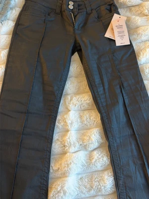 Svart coateda jeans från Nelly - Snygga svarta coateda jeans från Nelly med utsvängda ben och hög midja. Jeansen har två silverfärgade knappar och dragkedja framtill samt bälteshällor. Materialet har en lätt glansig finish som ger en trendig look.