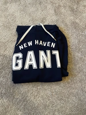 Mörkblå hoodie från GANT - Mörkblå hoodie från GANT med vit text 'NEW HAVEN GANT' på bröstet. Klassisk modell med huva och vita dragsnören. Mjuk bomullsblandning och ribbade muddar vid ärmslut och nederkant. Perfekt för en avslappnad och stilren look. Säljer denna hoodie för att jag växte ur den innan jag hann använda den särskilt mycket. 