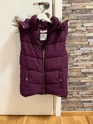 Lila dunväst med fuskpäls från H&M - Snygg lila dunväst från H&M med avtagbar huva och fuskpälsdetalj. Västen har dragkedja framtill, två fickor med tryckknappar och quiltad design. Perfekt för lager-på-lager och håller dig varm under kyliga dagar.