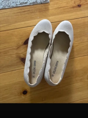 Beige ballerinaskor från Gyllene Gripen - Snygga beige ballerinaskor från Gyllene Gripen med vågig kant runt öppningen. Skorna har en klassisk rund tå och är tillverkade i ett mjukt material som liknar mocka. Perfekta för en stilren och enkel look.