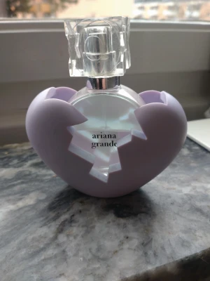 Ariana Grande parfym med lila hjärtskal - Lite mer än hälften kvar,se bild. Säljer en parfym från Ariana Grande med en unik rund glasflaska och ett transparent, geometriskt lock. Flaskan kommer med ett lila hjärtformat skal som kan tas av. Perfekt för dig som gillar snygga och kreativa parfymflaskor.