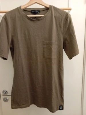 Olivgrön t-shirt med ficka från LMTD - Olivgrön t-shirt från LMTD med rund halsringning och en bröstficka. Tillverkad i mjuk bomull och har en stilren, enkel design med korta ärmar. Perfekt för en avslappnad och trendig stil.