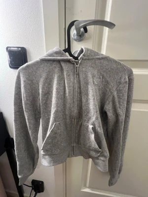 Grå hoodie från Juicy Couture - Säljer en grå hoodie från Juicy Couture med dragkedja och två fickor framtill. Hoodien har en broderad logga på bröstet och är tillverkad i mjukt material som känns riktigt skönt. Perfekt för dig som gillar en avslappnad och trendig look.