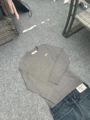 Hollister grå stickad tröja M - Grå stickad tröja från Hollister i mycket bra skick. Klassisk modell med rund halsring och en snygg neonfärgad logo på bröstet som ger lite extra detalj.  Storlek M med skön och normal passform. Perfekt basic-plagg som funkar till allt – jeans, cargos eller över skjorta.  Material: bomull (mjuk och bekväm) Skick: mycket bra  Passar perfekt för dig som gillar clean, casual eller streetwear stil.
