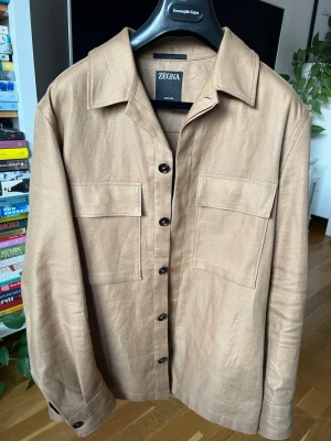 Zegna Oasi Lino Jacka/Overshirt RRP: 18000SEK - overshirt från Ermenegildo Zegna i den eftertraktade Oasi Lino-kollektionen – en sann representation av varumärkets kompromisslösa syn på kvalitet, innovation och hantverk. Plagget är tillverkat i 100% premiumlinne, framtaget från ZEGNAs egen Oasi där varje fiber är spårbar och noggrant utvald för att uppnå en överlägsen mjukhet, lätthet och andningsförmåga.  Längd: 71cm, bredd: 55, axelbredd: 46cm, längd ytter ärm: 65cm. Skickas med original galge och klädforal, RRP 20000SEK. Loro piana 