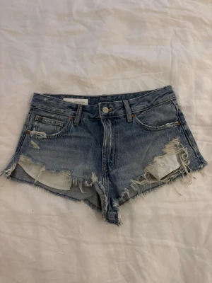 Blå slitna jeansshorts med rå kant - Säljer ett par blå jeansshorts med rå och fransig kant, slitna detaljer och klassisk femficksdesign. Shortsen har knapp och dragkedja framtill samt låg midja. Perfekta för sommarens chill dagar.