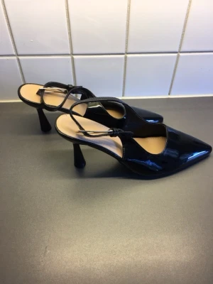 Svarta klackskor med slingback - ALDRIG ANVÄNDA! Snygga svarta klackar i lackmaterial med spetsig tå och slingback-rem. Klacken är hög och smal, vilket ger ett elegant intryck. Innersulan är ljusbeige och skon har en stilren design som passar till många olika outfits.