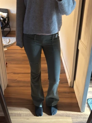 Gröna bootcut byxor med fickdetalj - Snygga gröna byxor med bootcut-snitt och cool fickdetalj på ena benet. Byxorna har två knappar i midjan och raka fickor framtill. Perfekta för dig som vill ha en trendig och bekväm look. 