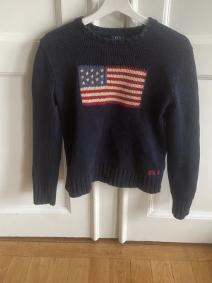 Mörkblå stickad tröja Polo Ralph Lauren - Mörkblå stickad tröja från Polo Ralph Lauren med amerikansk flagga på bröstet. Tröjan har rund halsringning, långa ärmar och ribbstickade muddar. Materialet ser ut att vara bomull och RL-broderi finns vid nederkanten.