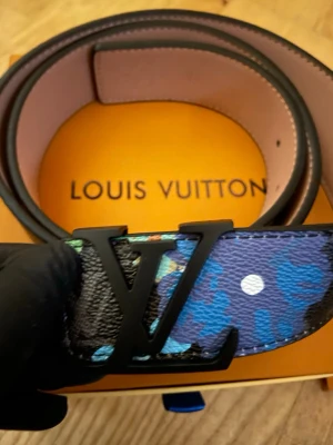  bälte med svart spänne - Snyggt bälte med stort svart spänne. Bältet har ett färgstarkt mönster i blått, svart, vitt och inslag av grönt och gult på präglat skinn. Perfekt accessoar för att lyfta din outfit.