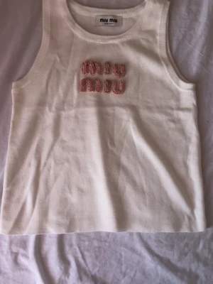 Vit ribbad linnetopp från Miu Miu - Supersnygg vit ribbad linnetopp från Miu Miu med rosa broderad logga framtill, dekorerad med små glittriga stenar. Perfekt att styla till jeans eller kjol för en trendig look.