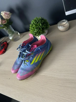 Blåa Adidas fotbollsskor med neon - Säljer ett par snygga fotbollsskor från Adidas i blått med neon-gula detaljer och rosa insida. Skorna har klassisk snörning och tålig syntetisk ovandel. Perfekta för dig som vill sticka ut på planen med både stil och funktion.