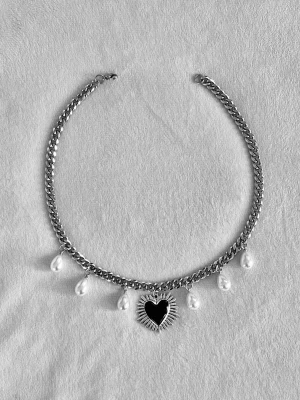 Silverfärgat halsband med pärlor och hjärta - Unikt halsband i silverfärgad rostfritt med droppformade vita pärlor och ett svart hjärthänge med solstråleliknande kant. Kedjan är chunky och ger en cool kontrast till de eleganta pärlorna. Perfekt för dig som gillar statement-smycken och vill sticka ut.