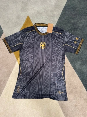 Svart och guld Nike fotbollströja - Cool svart fotbollströja från Nike med guldiga detaljer och geometriskt mönster. Tröjan har korta ärmar, rund hals och ett guldigt emblem på bröstet. Materialet är lätt och andas, perfekt för träning eller match.