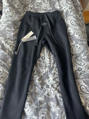 Svarta Essentials mjukisbyxor - Svarta mjukisbyxor från Fear of God Essentials med dragsko i midjan och diskret logga framtill. Byxorna har en avslappnad passform och är tillverkade i mjukt bomullsmaterial. Perfekta för en chill och stilren look.