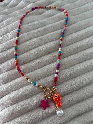 Färgglatt pärlhalsband med berlocker - Unikt halsband med små färgglada pärlor i olika nyanser och guldtonade detaljer. Halsbandet har tre berlocker: en lila sjöstjärna, en orange fisk och en vit pärla. Låset är i guldfärg och sitter framtill för en trendig look. Perfekt för dig som gillar lekfulla accessoarer.